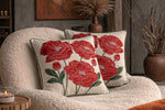 Two square throw cushion covers with red poppy embroidery on a cream background, styled on a textured beige armchair in a cozy room setting.